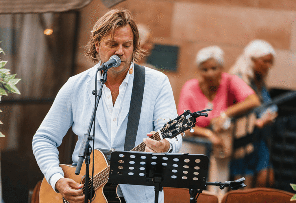 En man i ljusblå kavaj spelar gitarr utomhus.