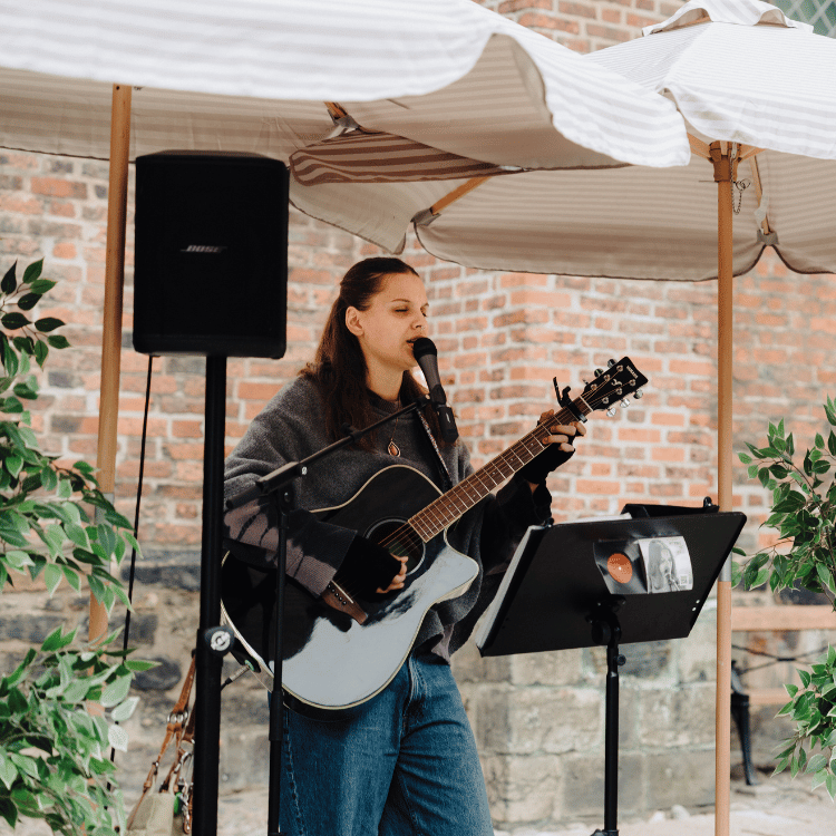 En tjej spelar gitarr och sjunger i en mikrofon. Bakom henne står en högtalare och två parasoll.