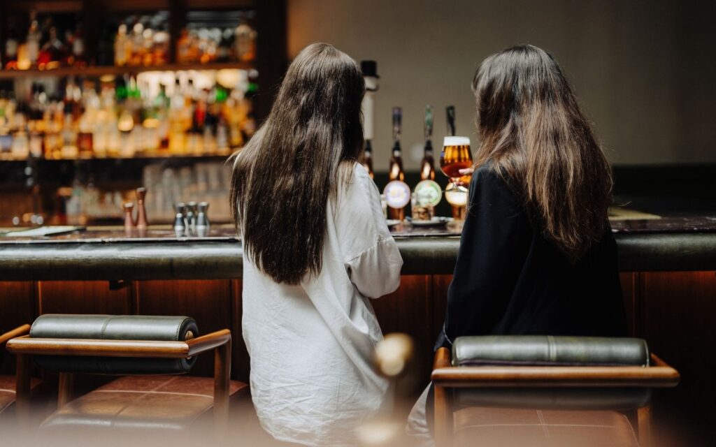 Två kvinnor står i en bar och den ena håller upp ett glas med öl