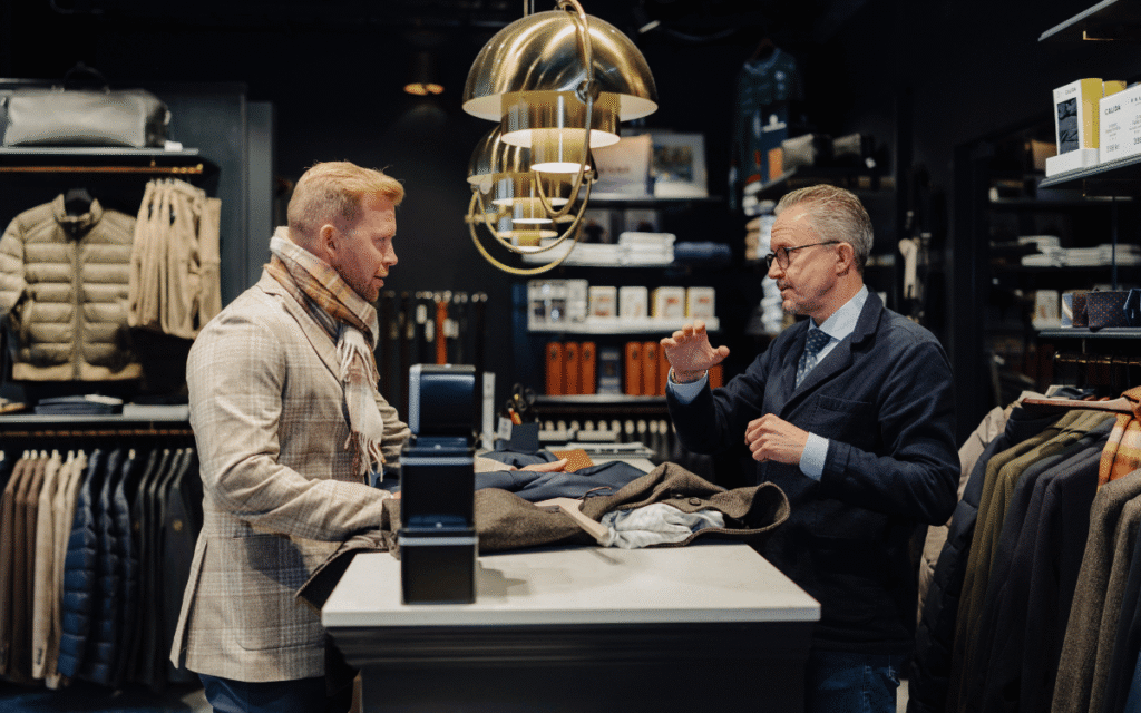 En man i jacka och halsduk står i kassan i en butik och handlar av en man i kostym.