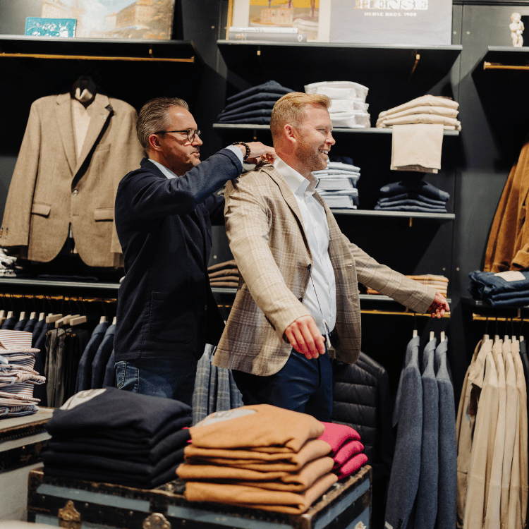 En man i kostym hjälper en annan man prova en kavaj i en butik.