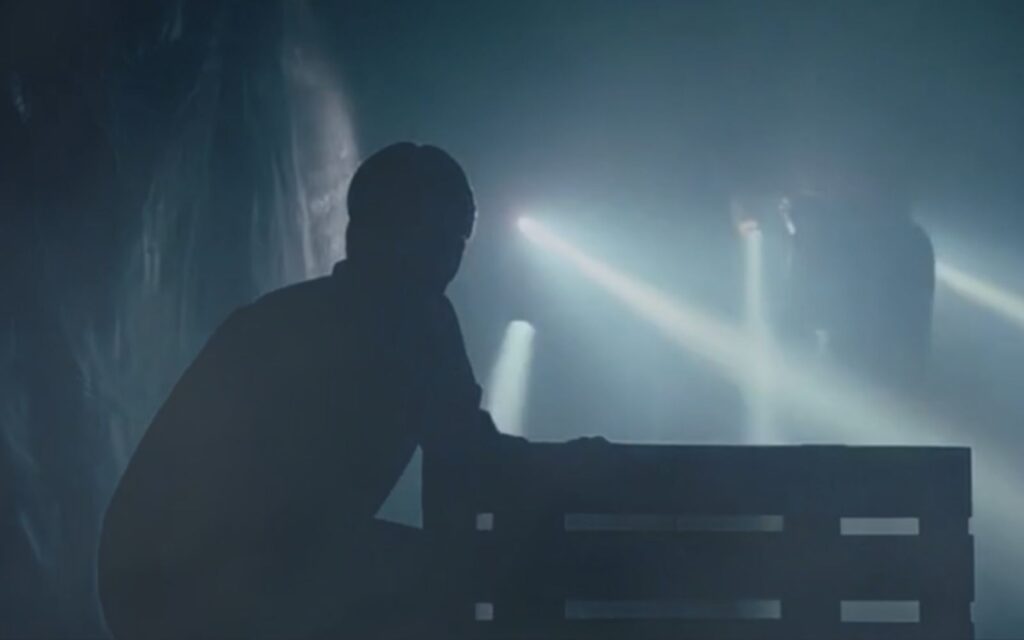 Ett mörkt rum, ser en gestalt som gömmer sig och i bakgrunden är et personer med ficklampa
