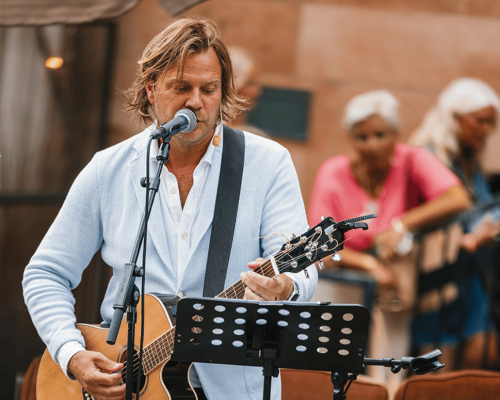 En man i ljusblå kavaj spelar gitarr utomhus.
